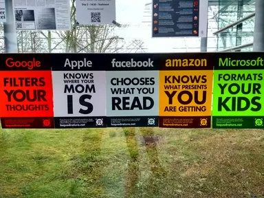 Five colorful posters displayed on a grassy area in front of a modern glass-and-metal building. From left to right: A red Google poster reading 'FILTERS YOUR THOUGHTS', a white Apple poster with 'KNOWS WHERE YOUR MOM IS', a gray Facebook poster stating 'CHOOSES WHAT YOU READ', a yellow Amazon poster with 'KNOWS WHAT PRESENTS YOU ARE GETTING', and a green Microsoft poster reading 'FORMATS YOUR KIDS'. All posters feature the π symbol and reference the website laquadrature.net. In the background, metal structures and leafless trees are visible.
