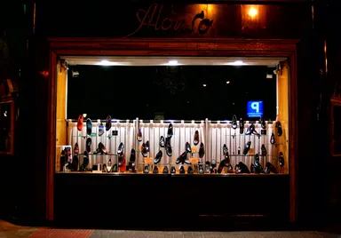 A nighttime storefront window display of a shoe shop named 'Alonso,' showcasing an array of men's dress shoes neatly arranged on glass shelves. The warm interior lighting highlights the polished leather shoes, while the dark exterior reflects the quiet street.