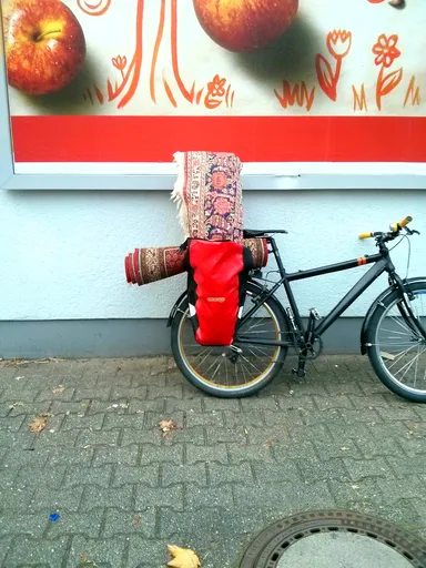 A black bicycle parked on a brick-paved sidewalk, leaning against a light gray wall. The bike is equipped with a red waterproof pannier bag on the rear rack and a rolled-up, intricately patterned red and beige carpet strapped horizontally across the handlebars. Above the bike, a large advertisement features illustrations of red apples and orange flowers on a white and red background. A manhole cover and scattered leaves are visible on the ground.