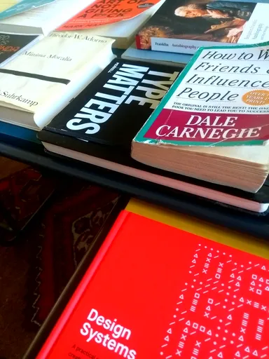 A stack of books on a couch table, including titles like 'How to Win Friends and Influence People' by Dale Carnegie, 'Type Matters!', and 'Design Systems'. The books are arranged in a slightly overlapping manner, showcasing their colorful covers.