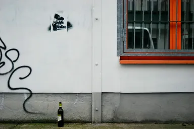 A bottle of wine standing on the ground in front of a white wall with graffiti and a sticker. To the right, a window with orange framing and metal bars is visible, reflecting a parked car outside.