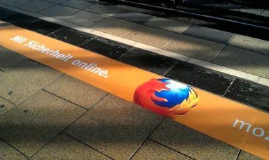 A close-up of an orange floor advertisement on a tiled pavement. The ad features the Firefox browser logo (a stylized fox wrapped around a blue globe) and the German text 'Mit Sicherheit online.' and 'moz://a' (partial Mozilla branding). The ad is placed between the tiles and the metal tram tracks, with the pavement stones visible in the foreground and background.