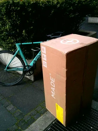 A light green Aventon road bike leaning against a concrete planter with dense green hedges in the background. Next to the bike, a large cardboard box labeled 'MADE' is placed on a paved sidewalk. The box has a circular symbol on top and a yellow shipping label on the side. The scene is outdoors, with the bike and box casting shadows on the pavement.