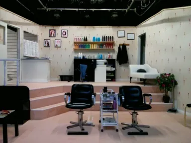 A theater stage showing a well-lit hair salon interior with two black salon chairs in the foreground, a rolling trolley with hair products. The walls are decorated with framed photos, a shelf of colorful hair products, and a white armchair. A small potted plant adds a touch of greenery.