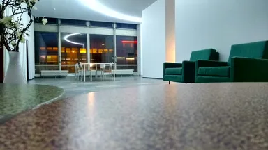 A modern, well-lit lounge area with two green armchairs in the foreground from the 50s. In the background, a glass-walled space features a white dining table and chairs, with a view of city lights at dusk. A tall white vase with decorative branches adds a touch of elegance to the room.