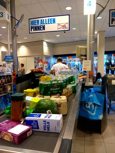 A supermarket checkout area with a conveyor belt filled with groceries, including beer packs, a watermelon, a jar of coffee, and a carton of whole milk. A sign above reads 'HIER ALLEEN PINNEN' (Only Card Payments Here). Customers and cashiers are visible in the background.