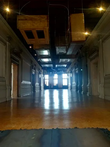 A long, dimly lit hallway in a Venetian villa, featuring ornate columns, high ceilings, and suspended wooden cabinets. The polished floor reflects the soft lighting, leading to a brightly lit room at the end.