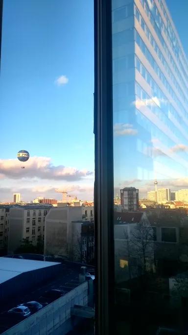 A cityscape view through a window, split by the window frame. In the distance, a white and blue airship with the text 'DIE WELT' floats in the sky above a mix of modern and older buildings. The Fernsehturm (TV Tower) and other high-rise buildings are visible under a partly cloudy sky with soft sunlight. Below, a parking lot with a few cars and bare trees is visible, framed by the window’s reflection.