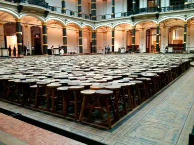A spacious, ornate indoor hall with high arched ceilings, dark columns with gold accents, and intricate tiled flooring. In the center, a large rectangular arrangement of rustic wooden stools with round seats fills the space. A few visitors are scattered around the edges of the hall, observing the installation. The lighting is warm, highlighting the architectural details and the contrast between the modern art installation and the historic setting.