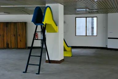 A bright yellow and blue slide structure with a ladder, positioned indoors against a white pillar. The slide is part of a minimalist, empty room with wooden doors, a window, and a concrete floor, creating a playful contrast with the industrial surroundings.